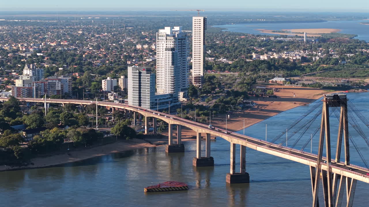 High-altitude drone tracking right, focusing on the bridge towers against the river and the vegetated cityscape of Corrientes