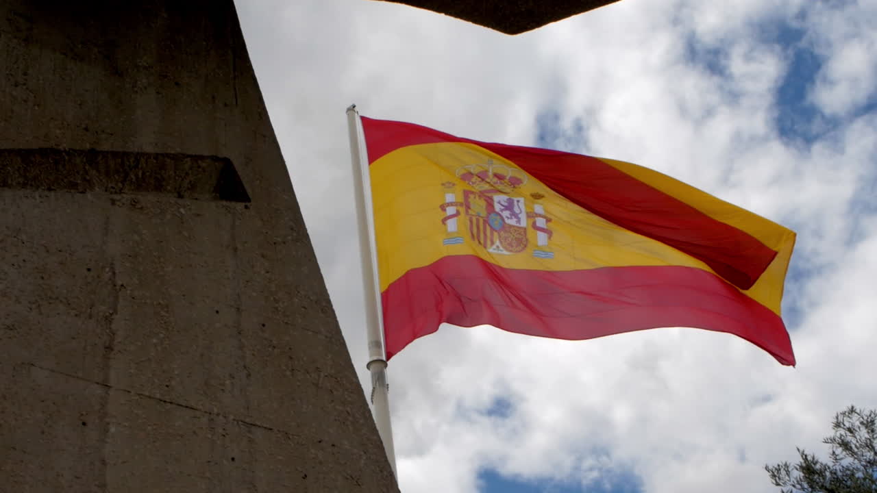 The Spanish flag waves proudly against a textured concrete structure and a sky filled with light clouds, evoking strength and identity.