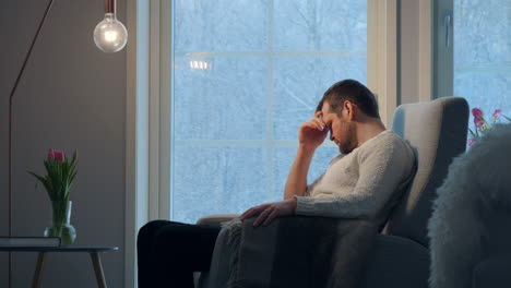 Young-man-sitting-sad-inside-modern-home-during-heavy-snowfall,-side-view