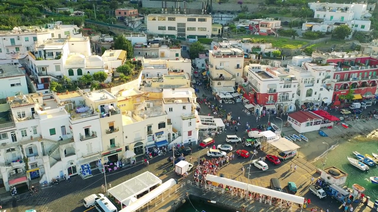 Ripresa aerea con drone di una città costiera in Grecia che mostra una piazza affollata con pedoni e veicoli e un porto con barche, circondata da edifici bianchi e colorati