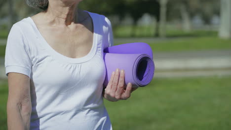 Middle-aged-female-body-walking-in-park-with-yoga-mat-in-hand