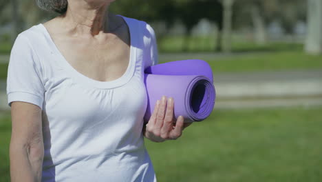 middle-aged female body walking in park with yoga mat in hand