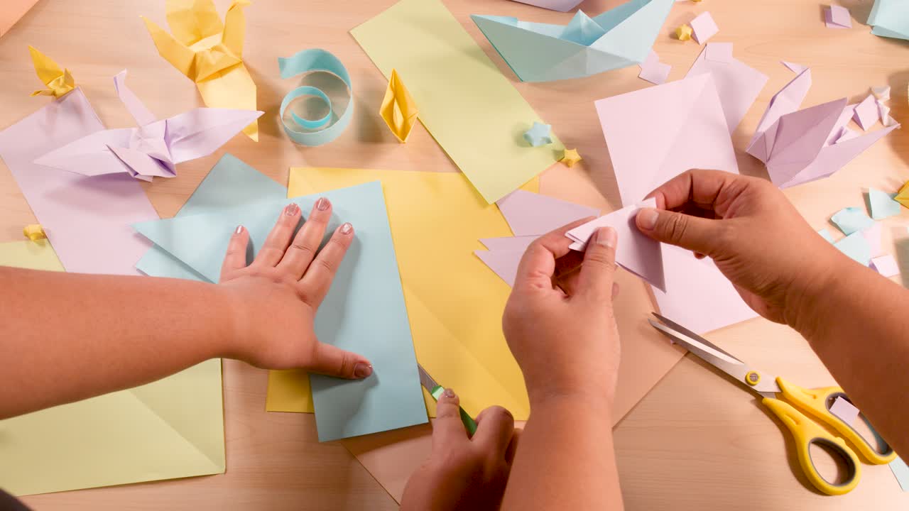 Two hands fold pastel origami paper on a well-lit desk with craft supplies, overhead view