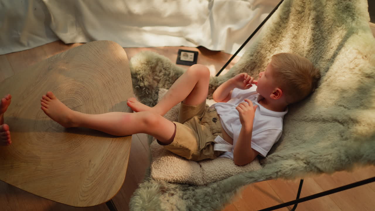 el niño se sienta en una silla de pelo en una mesa baja. el niño descalzo descansa en un sillón suave en el salón doméstico. el niño relajado disfruta de una mañana tranquila de fin de semana