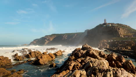 Waves-crash-over-reef-at-The-Poort,-lighthouse-on-mountain
