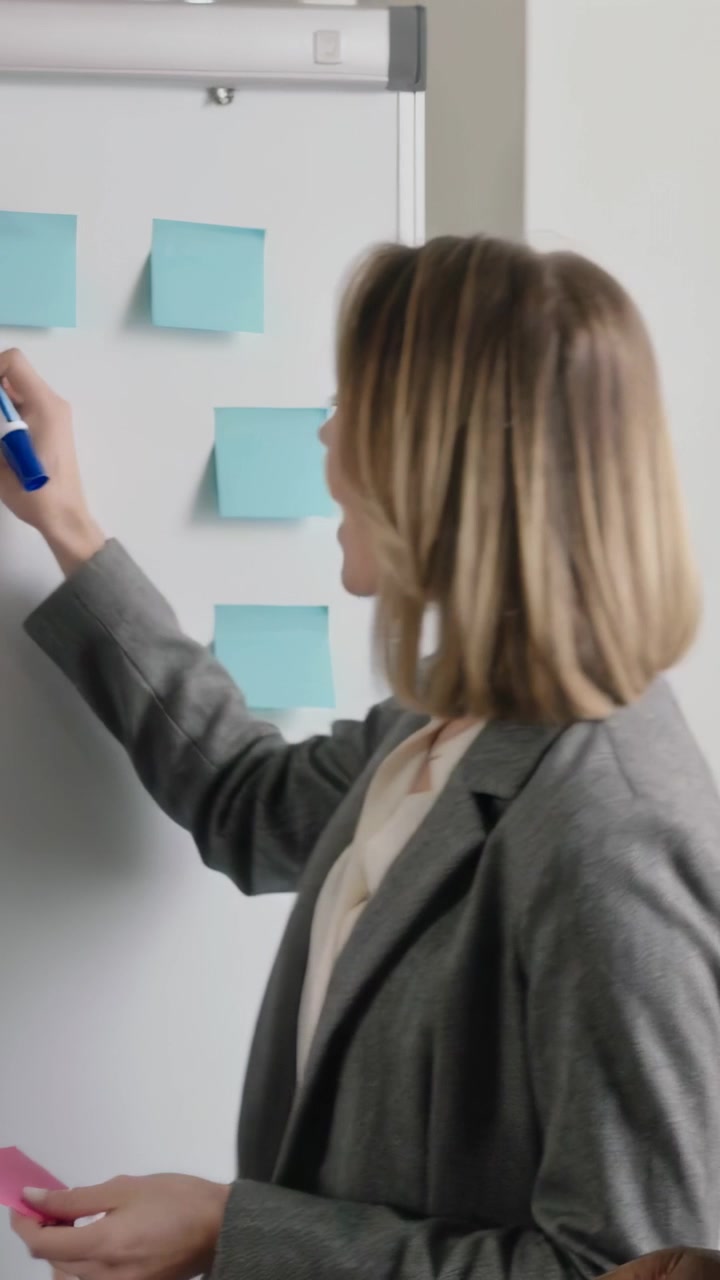 Video verticale: Mani che raggruppano appunti, donna in blazer grigio che scrive su una lavagna con un pennarello blu, uomo che si unisce.
