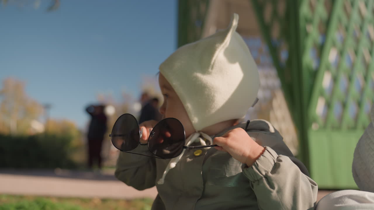 Bambino al parco che esplora occhiali da sole, berretto verde e cappotto caldo, luce solare autunnale, sagome di adulti sullo sfondo, gita familiare spontanea che cattura curiosità, gesti giocosi, sguardo quieto e relazione genitore-figlio.