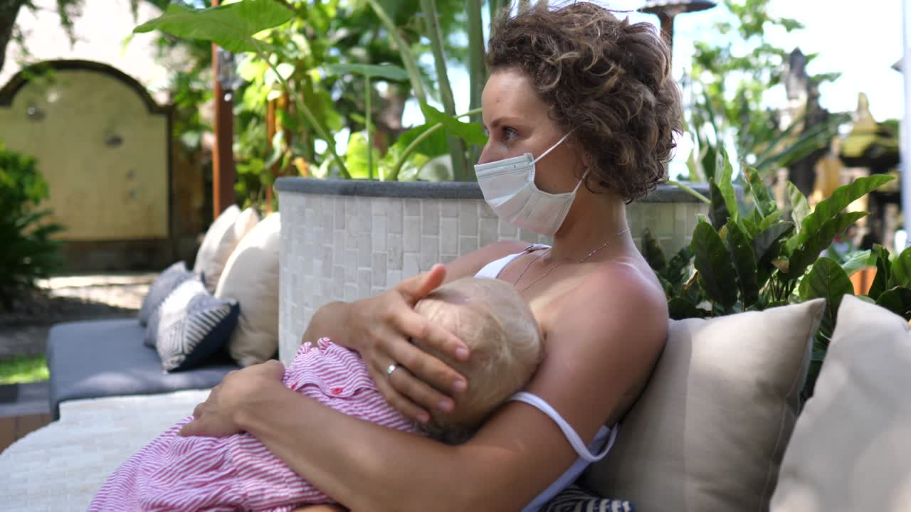 joven mujer caucásica con una máscara facial amamanta a su hija en un restaurante de jardín