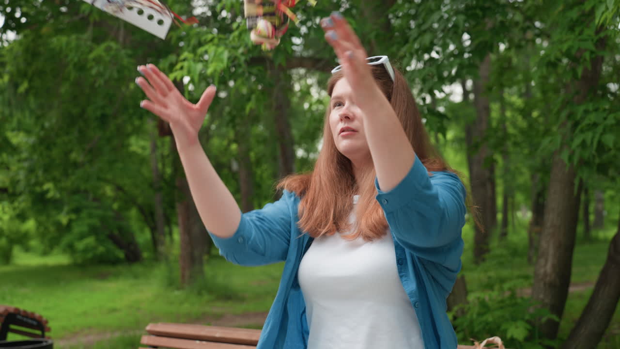 Молодая женщина на улице подбрасывает в воздух разноцветные нитки для вышивания, игриво двигаясь, в синей рубашке и белом наряде, сидя на деревянной скамейке в зеленом парке, выражая радость и творческую энергию