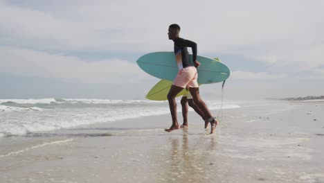 padre afroamericano y hijo adolescente corriendo en una playa con tablas de surf y hablando