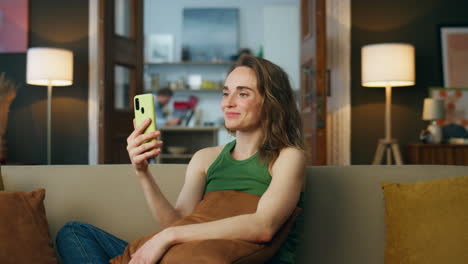 Friendly-lady-waving-hand-speaking-smartphone-home.-Relaxed-woman-sitting-sofa