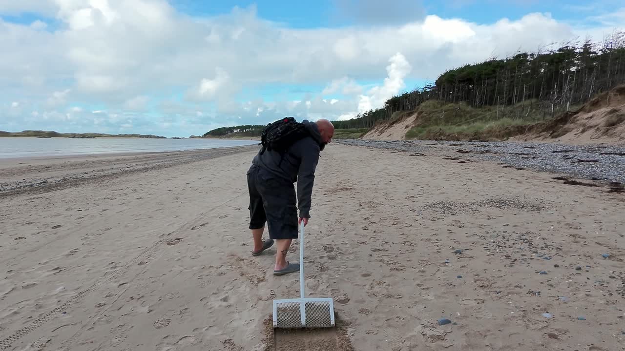 Bald male carrying sand scoop walking pet dog on sunny Welsh beach active lifestyle exploring