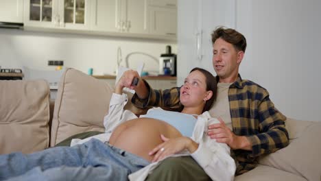 a man in a plaid shirt and his pregnant wife are relaxing on the sofa and laughing while watching tv at home