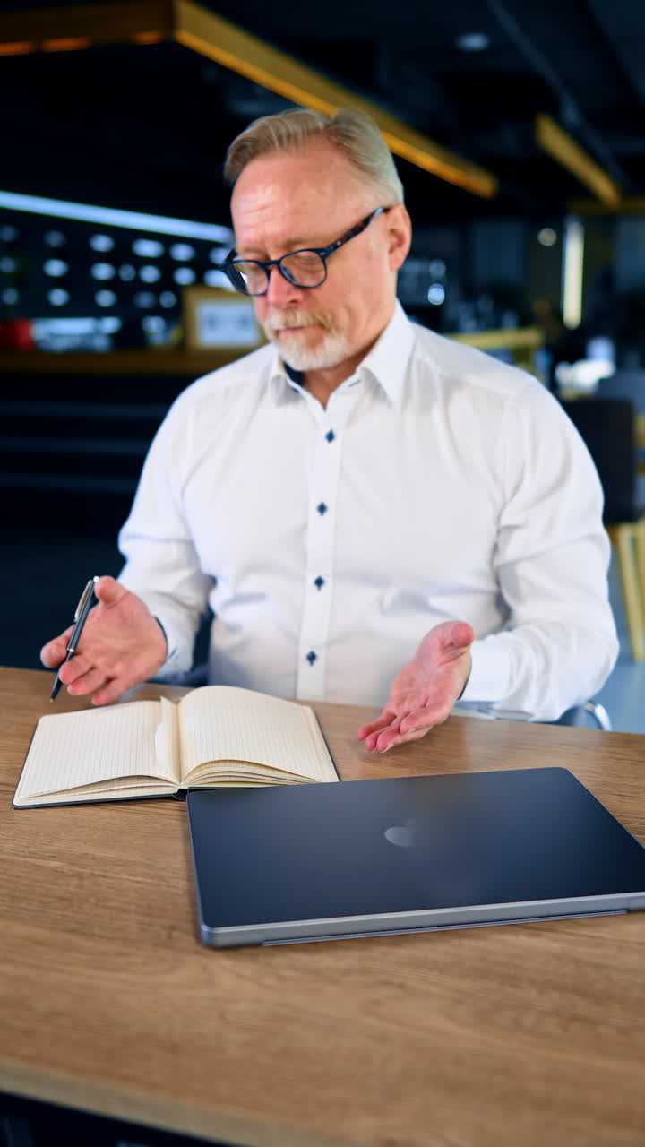 Bemused adult male wearing glasses and white shirt sits at desk in office. Man pulls the paper strip from his notebook and chews it. Vertical video.