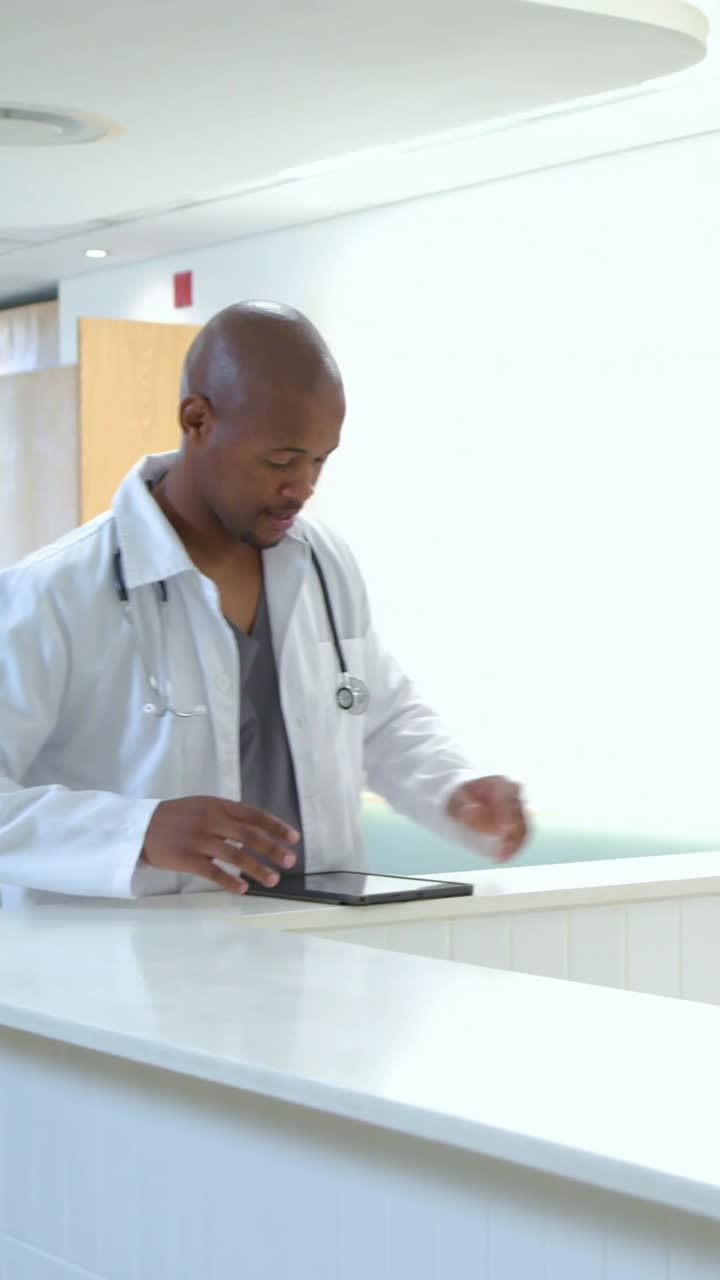 Video verticale: Medico con camice e stetoscopio che controlla un tablet alla reception, assistendo un paziente.