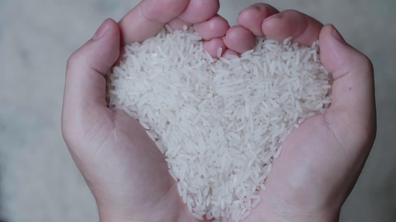 Hand Holding Raw Rice And Show Up In Slow Motion, Over White Rice Grains Background