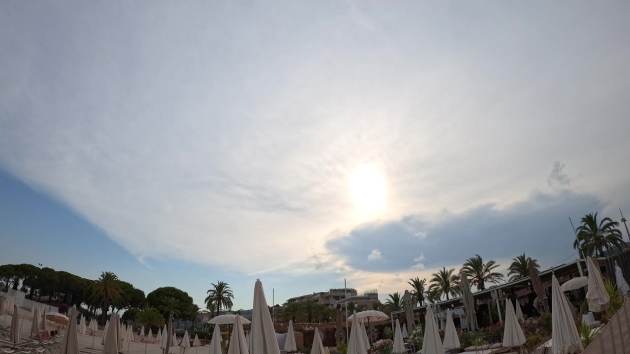 Resort sulla spiaggia con numerosi ombrelloni piegati, palme ed edifici contro un cielo nuvoloso con il sole che filtra