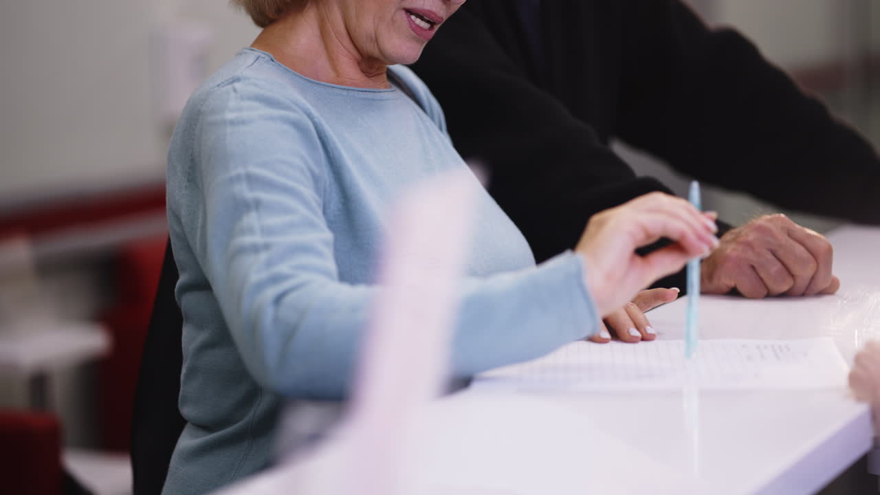 Senior woman writing at a desk with a man in the background