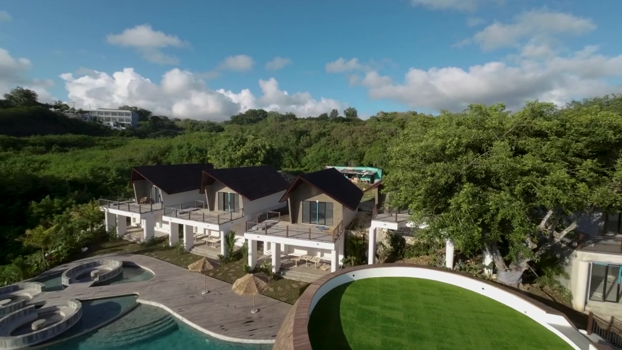 Toma aérea cinematográfica que muestra villas de resort modernas, una piscina única y un exuberante paisaje verde bajo un cielo azul brillante en Bali