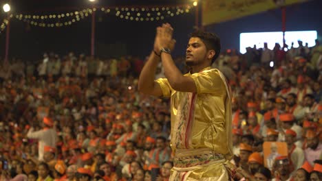 young priest performing ganga aarti rituals at holy hindu religious temple assi ghat in the evening with large crowd of people sitting in background, bjp political campaign public event