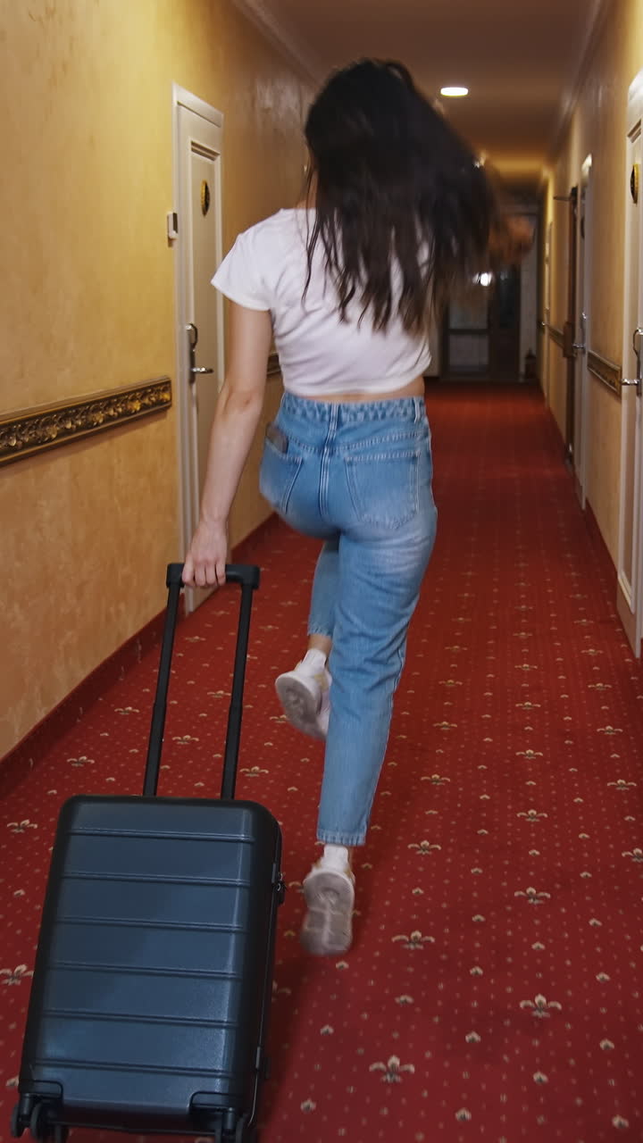 Happy female traveler with suitcase indoors. Young woman in casual clothes walking along the hotel corridor with trolley bag and jumping joyfully. Vacation concept. Vertical video