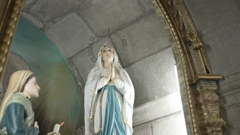 statue of the virgin mary in prayer with a blue and white robe, placed inside a church alcove with soft lighting