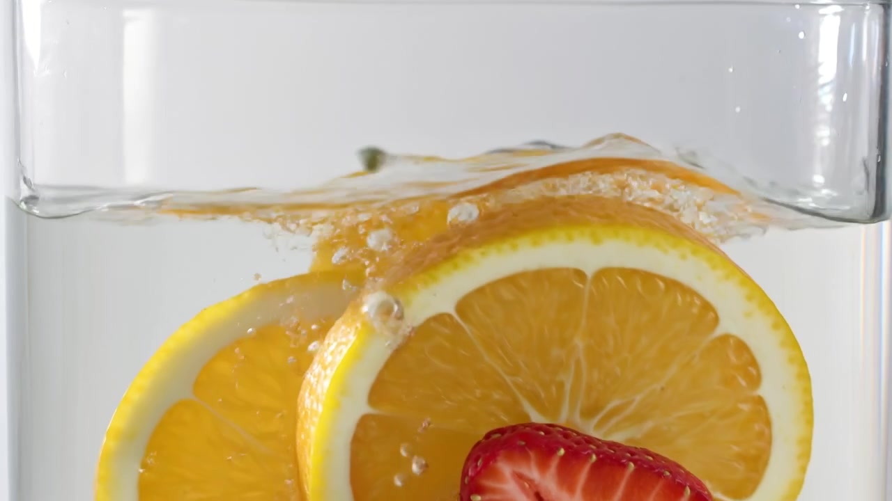 Fetta d'arancia fresca e fragole a fette che schizzano in acqua limpida in un contenitore di vetro trasparente.