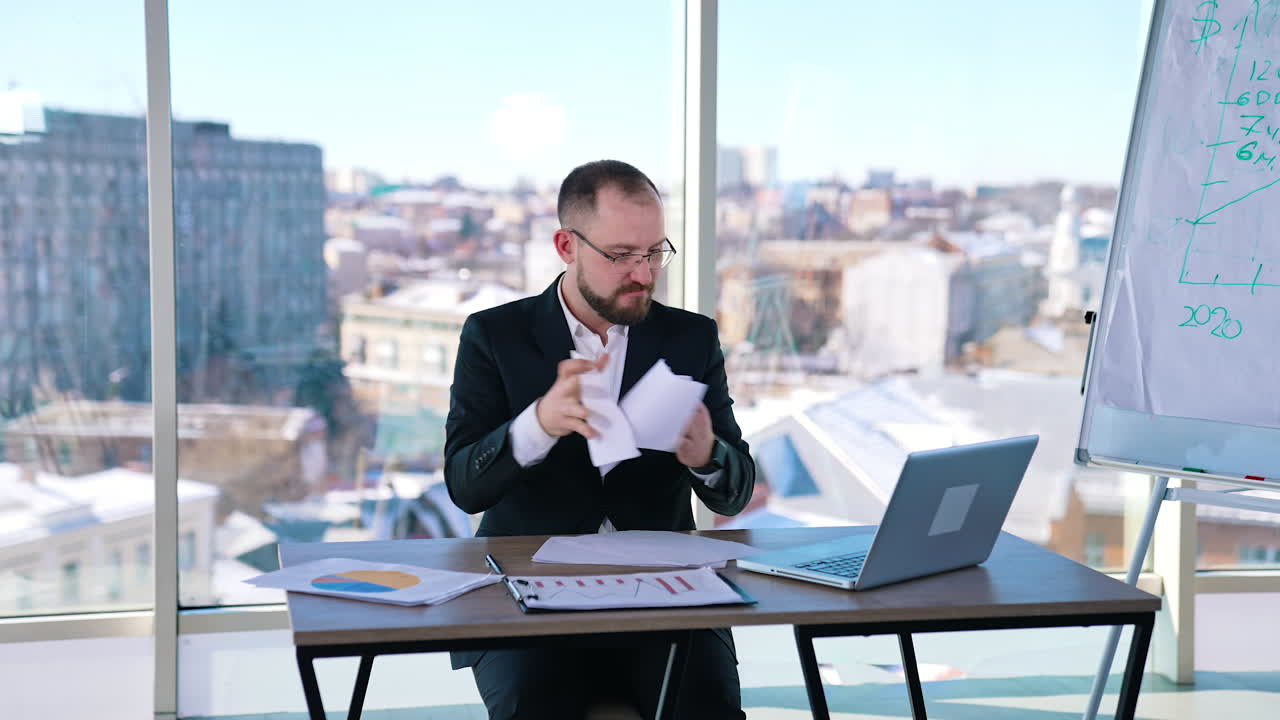 Nervous entrepreneur on his workplace. Businessman in costume sitting at the desk and throwing papers. Business problems.