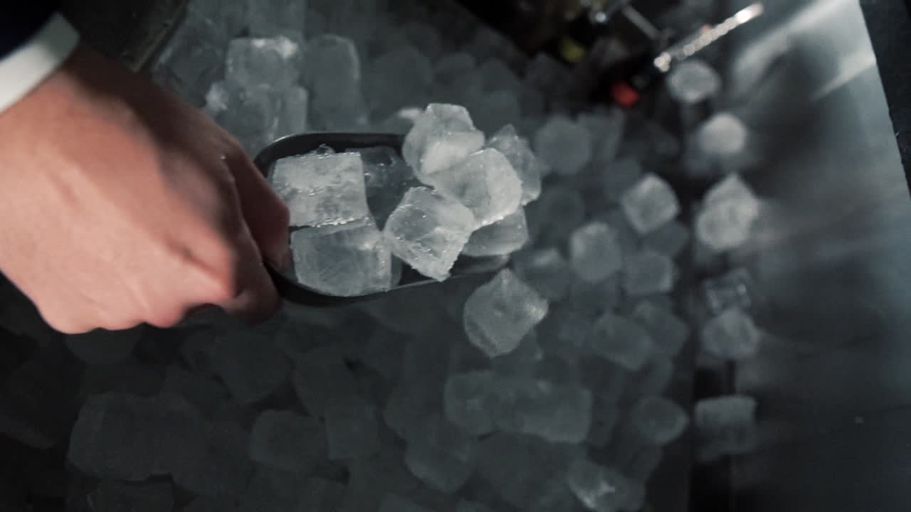 A bartender is firmly holding the ice scoop and place several ice cubes into a stirring glass. Dynamic video clip.
