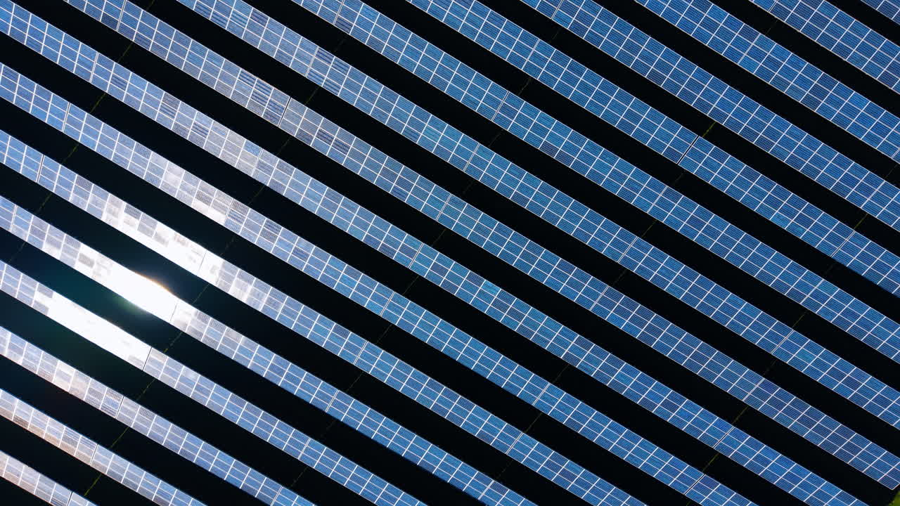 Vista aerea dall'alto di file di pannelli solari con riflesso del sole. Ripresa con drone di file di pannelli fotovoltaici su terreno scuro con la luce solare intensa che si riflette dalla superficie.