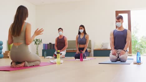 Instructora-Caucásica-Explicando-A-Un-Grupo-Diverso-Con-Máscaras-Faciales-Arrodilladas-Sobre-Colchonetas-En-Clase-De-Yoga