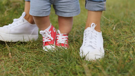 close up of an unrecognizable baby boy with his mother wearing sneakers and taking his first steps on the green grass