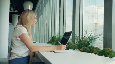 formal woman with laptop and papers. side view of businesswoman writing on paper while sitting with laptop at table near window in modern office