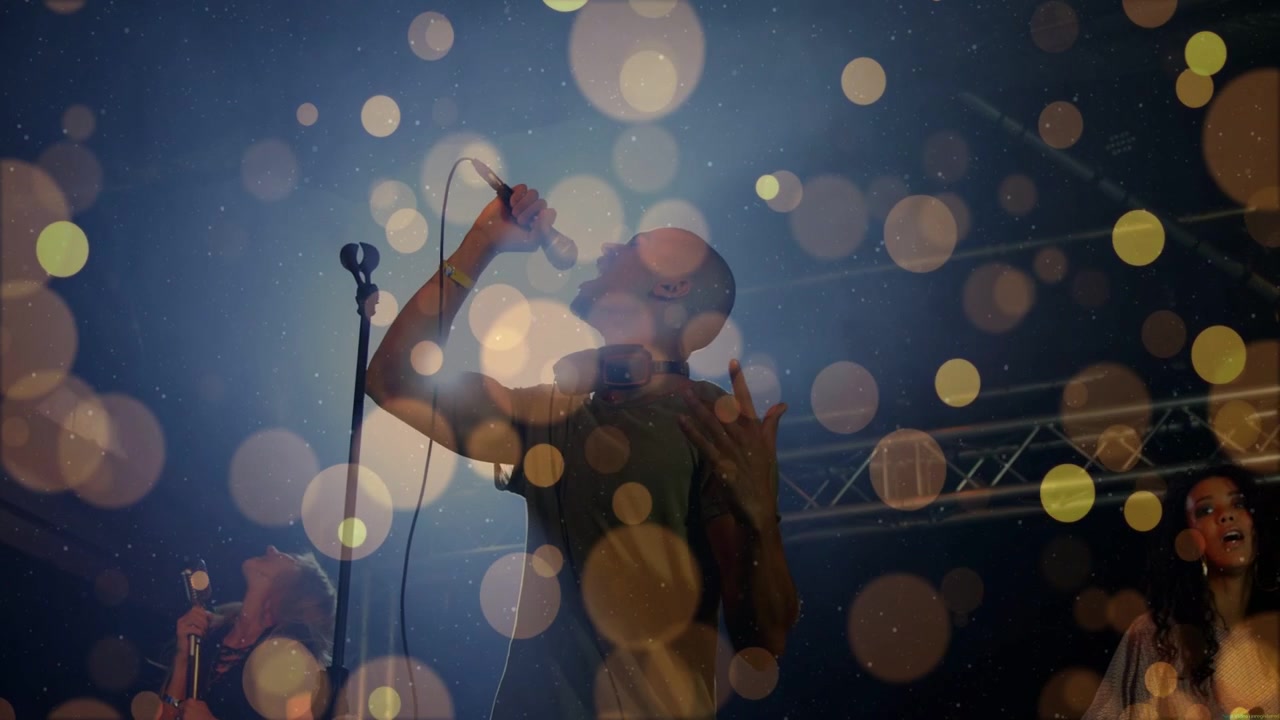 Cantante principal masculino cantando en un concierto con micrófono, bokeh apareciendo y luego desapareciendo, revelando a la banda para crear ambiente.