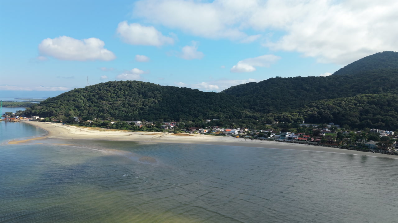 A beautiful aerial view of Prainha beach, located between Guaratuba and Matinhos in Paraná, Brazil. The drone captures the sandy peninsula, the calm bay, and lush green mountains.