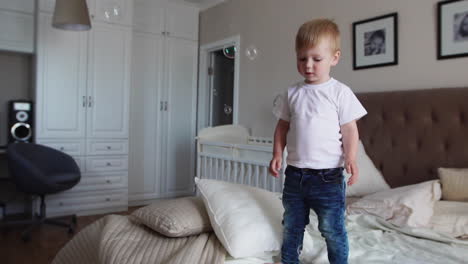 Ein-Junge-In-Einem-Weißen-T-Shirt-Und-Einer-Blauen-Jeans-Fängt-Seifenblasen-Ein,-Die-Auf-Dem-Bett-Im-Elternschlafzimmer-Stehen