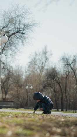 a young boy is seen squatting and digging the soil with a stick in his hand, he is dressed warmly in a black leather jacket, head warmer, and jeans, with trees and street lamps around