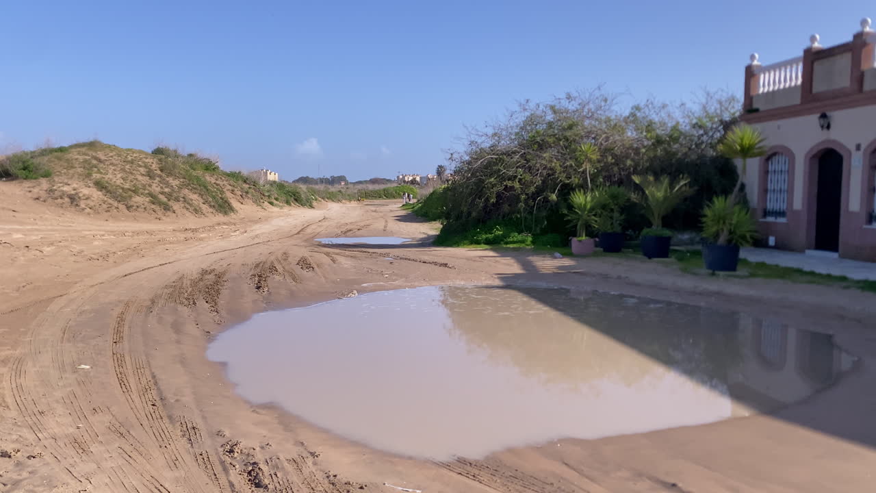 Sentiero sabbioso che riflette il cielo blu e una casa vicina, mostrando un ambiente costiero e terreno non asfaltato in Spagna
