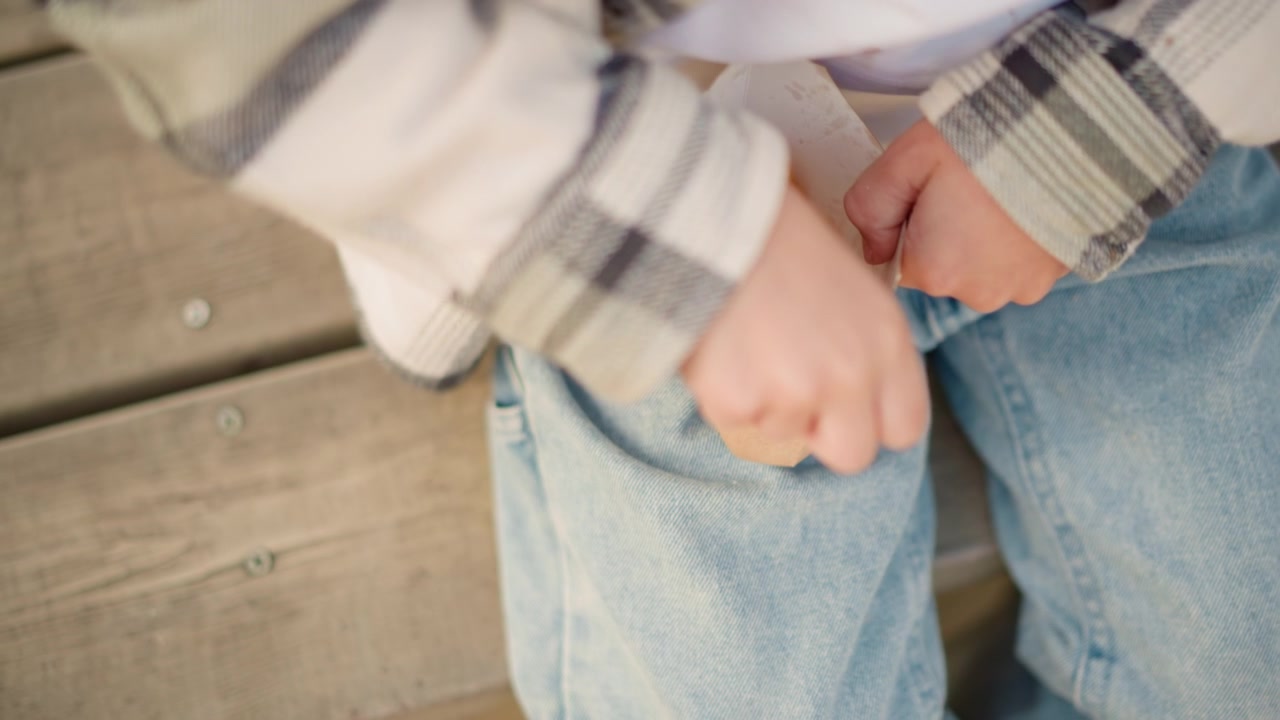 Primo piano di un bambino che tiene e cerca in un sacchetto di carta pieno di patatine fritte mentre è seduto su gradini di legno. Il bambino indossa jeans blu e una camicia a quadri concentrandosi unicamente sull'azione che coinvolge