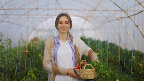 happy farmer woman holding basket with fresh harvested vegetables and smiling at greenhouse