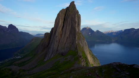 Dramatic-aerial-shot-of-Mountain-Segla-in-Senja-Norway,-with-a-tent-in-the-foreground-and-the-shear-rocky-cliff-face-drops-off-into-the-sea-with-the-craggy-mountain-range-in-the-background