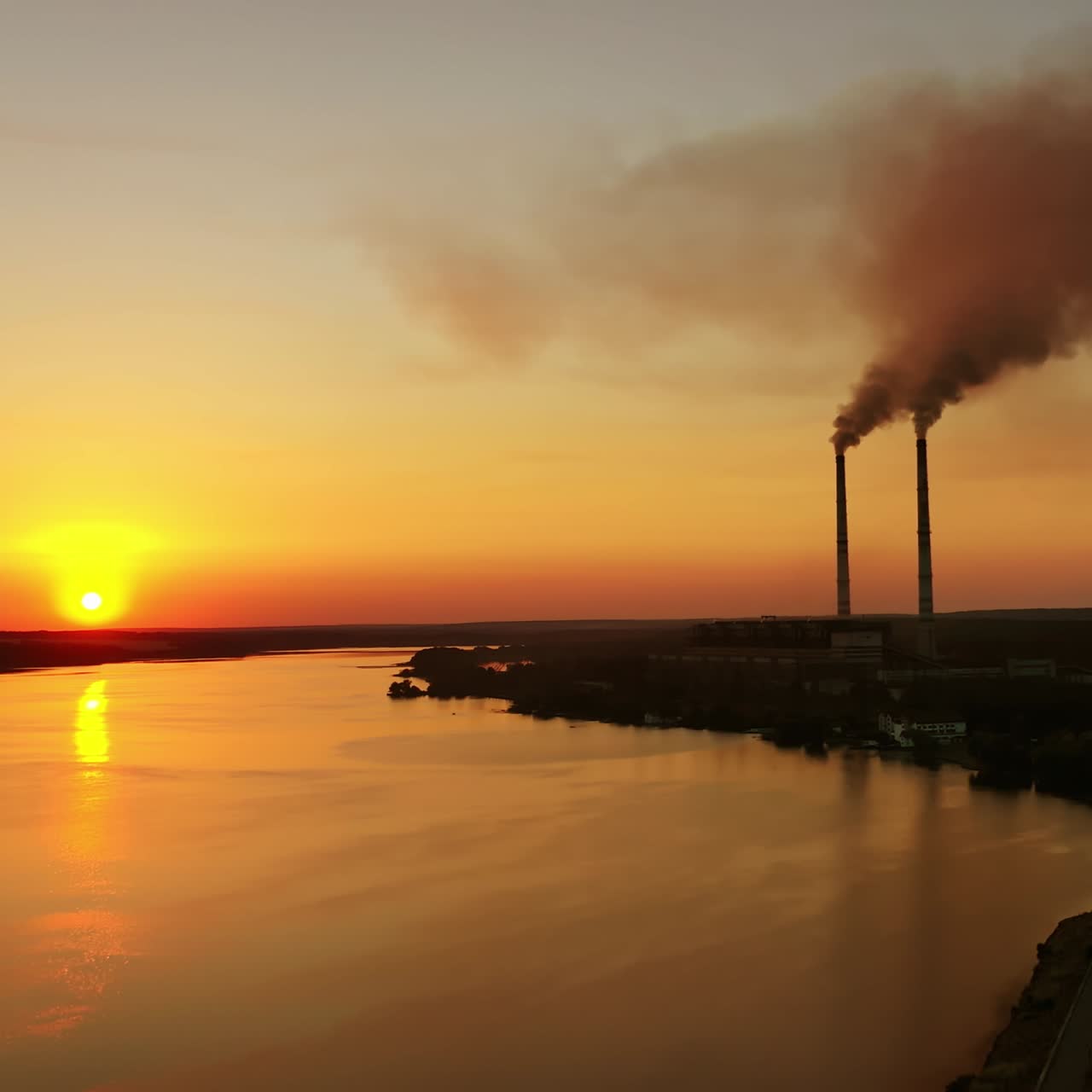 Industry near the river in the evening. Factory pipes release smoke into nature at sunset time. Pollution the environment. Aerial view. Camera moves back.