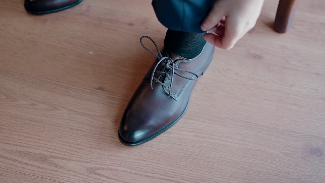 Close up of hands tying laces on a polished brown leather shoe on wood floor