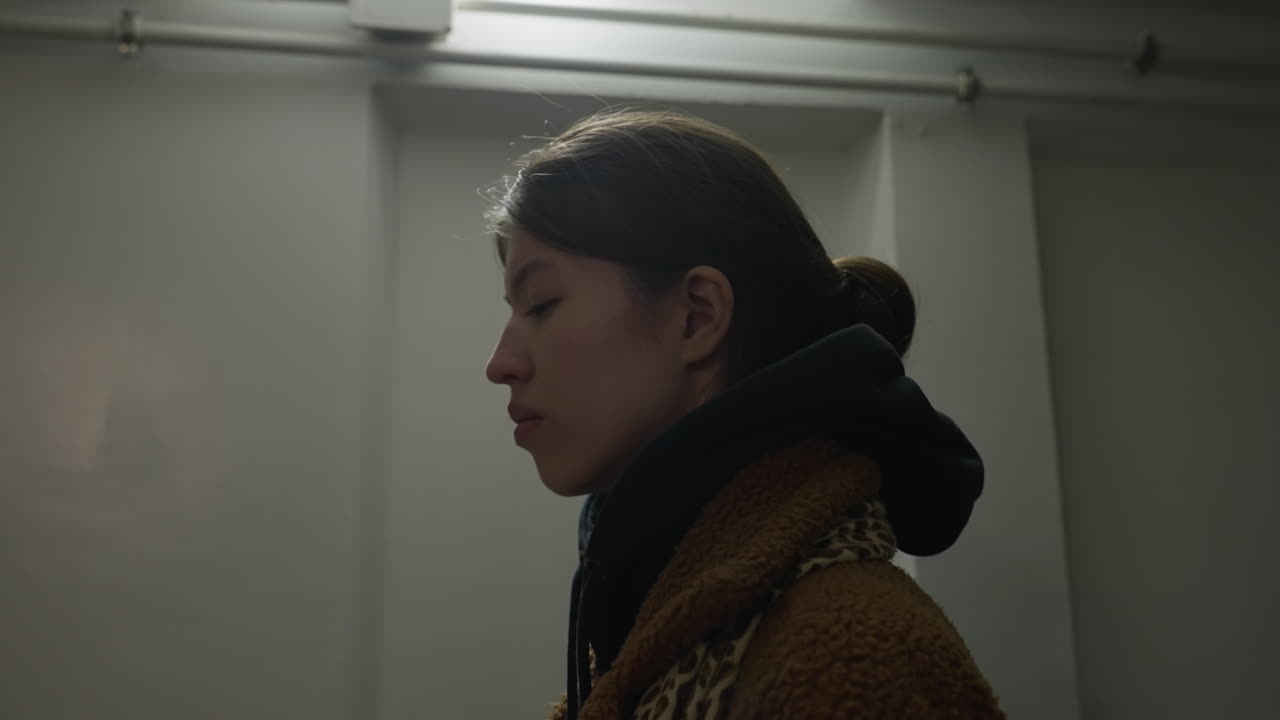 갈색 코트를 입은 괴로워하는 소녀의 클로즈업, 우울한 표정으로 올려다보며, 이 이미지는 미니멀하고 희미하게 조명된 환경에서 그녀의 감정의 강도를 포착합니다.