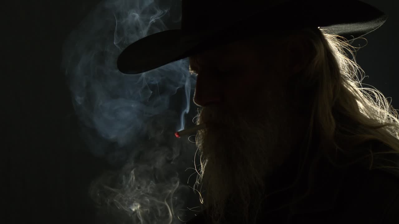 Backlit bearded man in Stetson hat lights cigarette, smoke curls