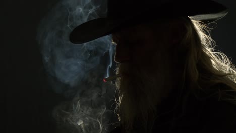 backlit bearded man in stetson hat lights cigarette, smoke curls