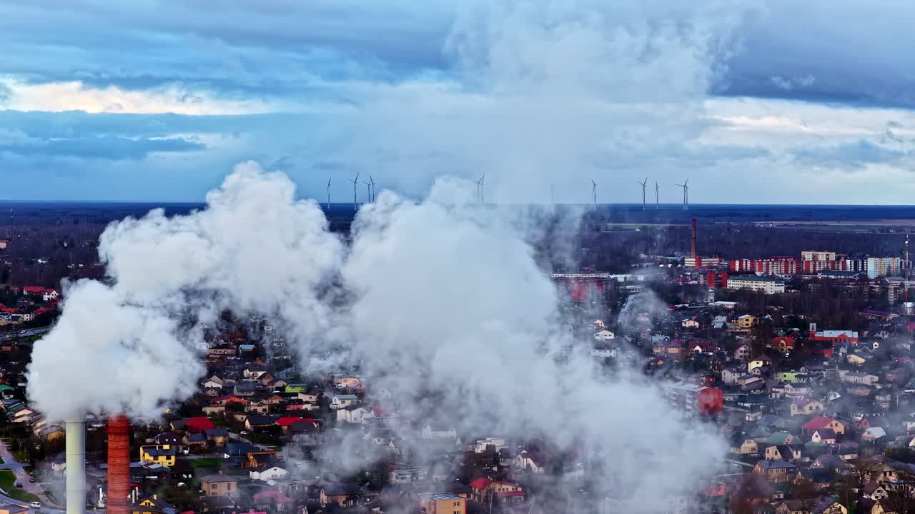 Vista aerea di una ciminiera industriale che rilascia vapore denso sui tetti di periferia, con turbine eoliche all'orizzonte sotto nuvole spettacolari