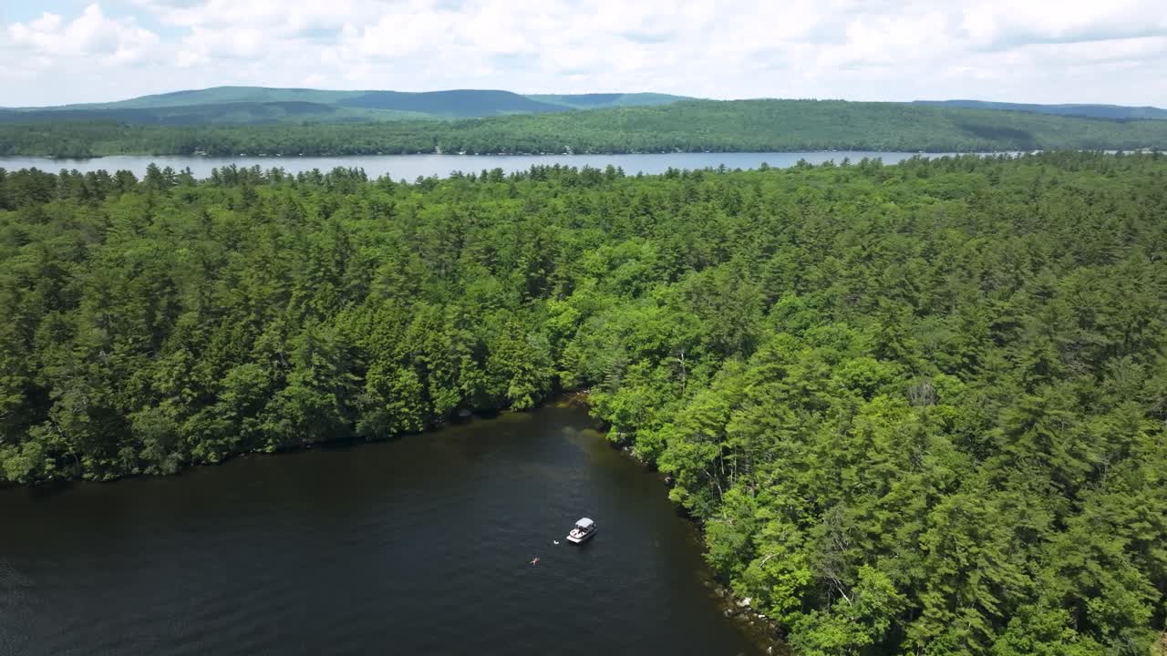 Aerial view of a pontoon boat anchored in a quiet cove on a forest-lined lake in Belgrade, Maine. A swimmer floats nearby, surrounded by summer greenery and distant hills.