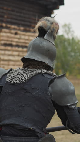 dark knight with skull on helmet swings spear rain. legendary medieval evil warrior fights enemies near building with burnt roof on cloudy day closeup