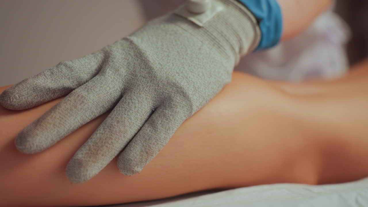 behansket hånd masserer klientens ben med mikrostrømshandske, ledende elektrodeplade på læg og ankel, blide cirkulære strøg fremmer cirkulation og afslapning, varm spa-belysning, klinisk og beroligende stemning